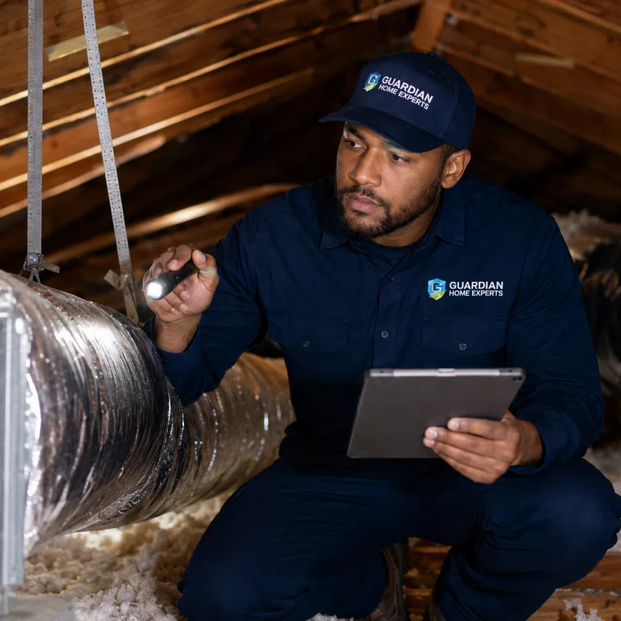 Technician in navy uniform inspecting attic HVAC ducts with flashlight and tablet under wooden roof beams.