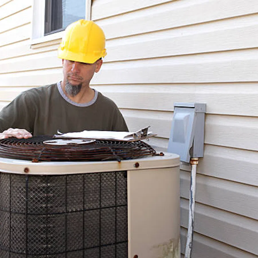 Technician in yellow hard hat inspecting outdoor air conditioning unit next to house siding with clipboard.