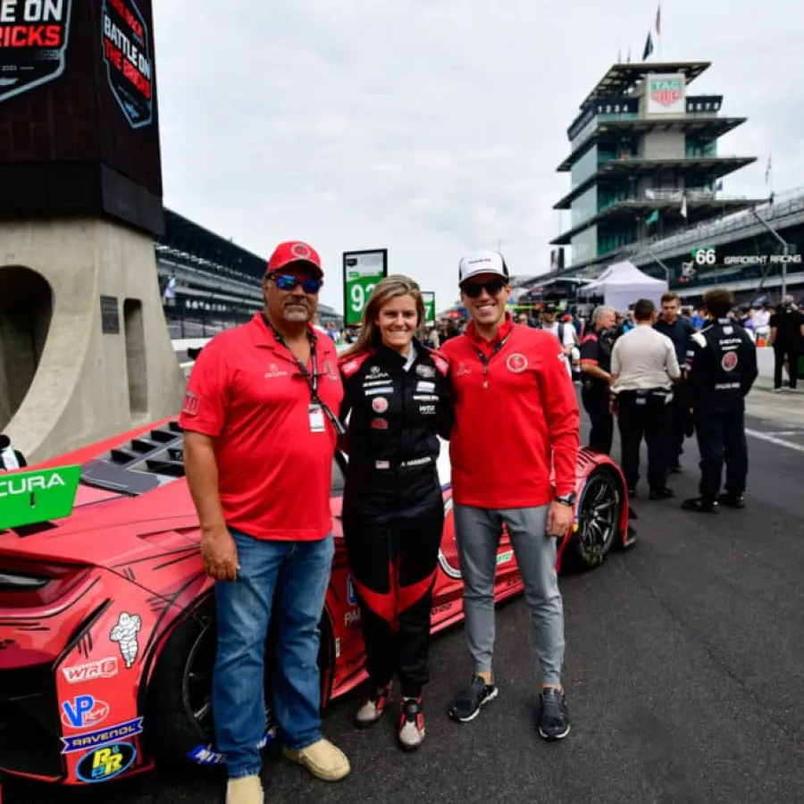 Three people posing next to a red Acura race car on a racetrack with banners and pit crews in the background.