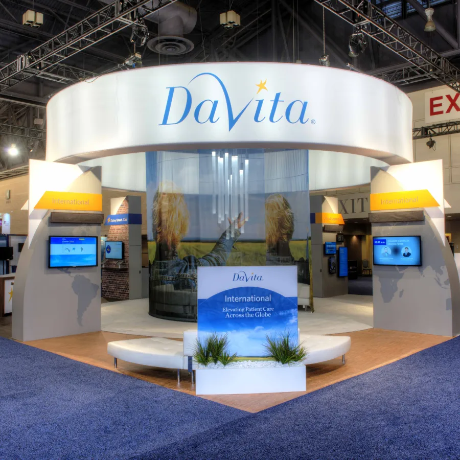 DaVita trade show booth with curved display and blue carpet at a convention center with exit signs.