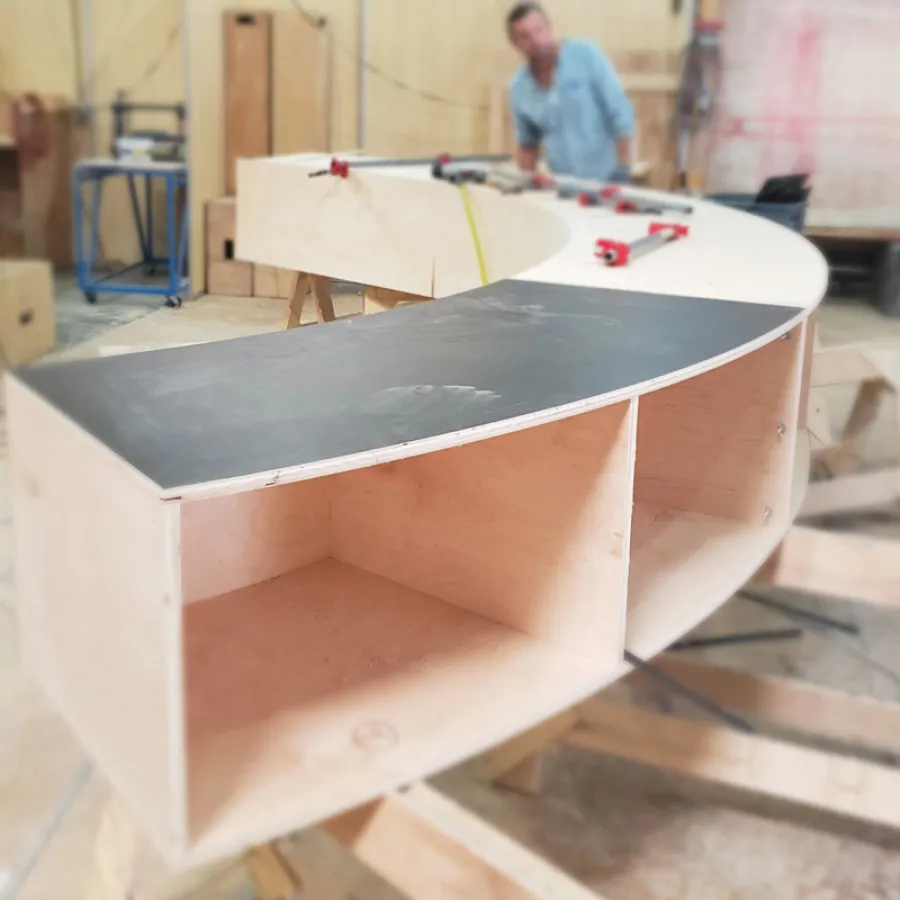 Curved wooden shelving unit under construction in a workshop with tools and blurred worker in background