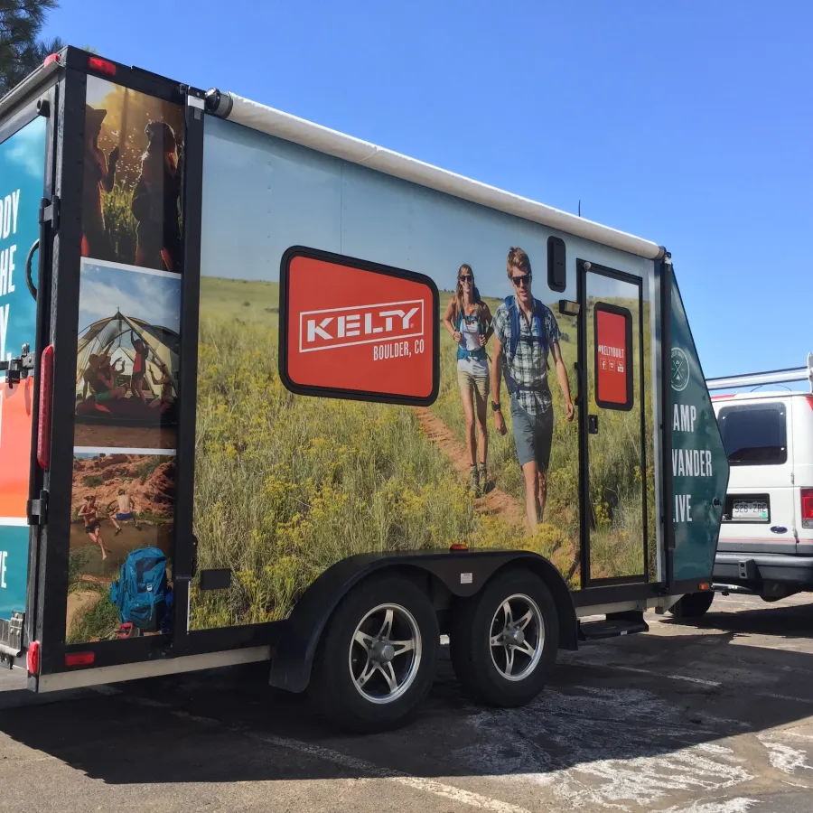 Kelty branded trailer and van with outdoor adventure scenes and slogan parked in a sunny lot.