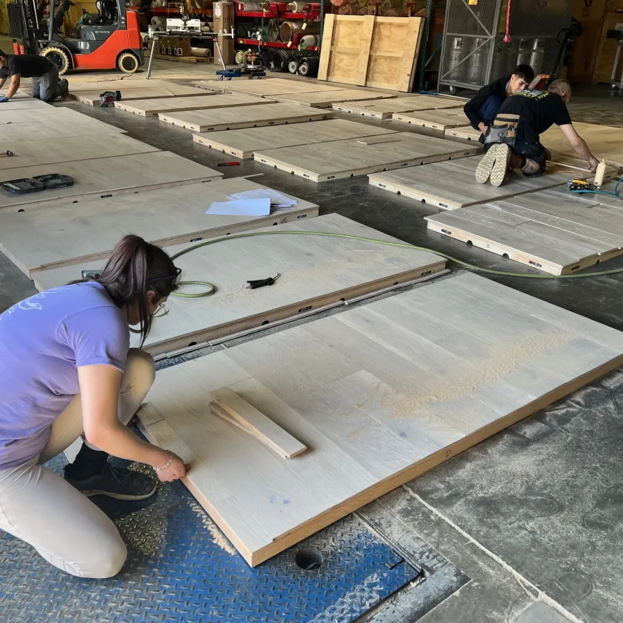 Workers assembling large wooden panels on a factory floor using various tools and measuring tapes.