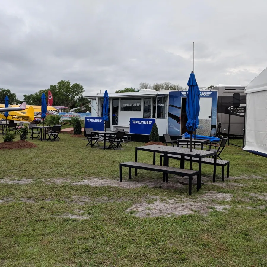 Outdoor seating area with black tables, blue umbrellas, and airplanes displayed near a Pilatus promotional trailer and tent.