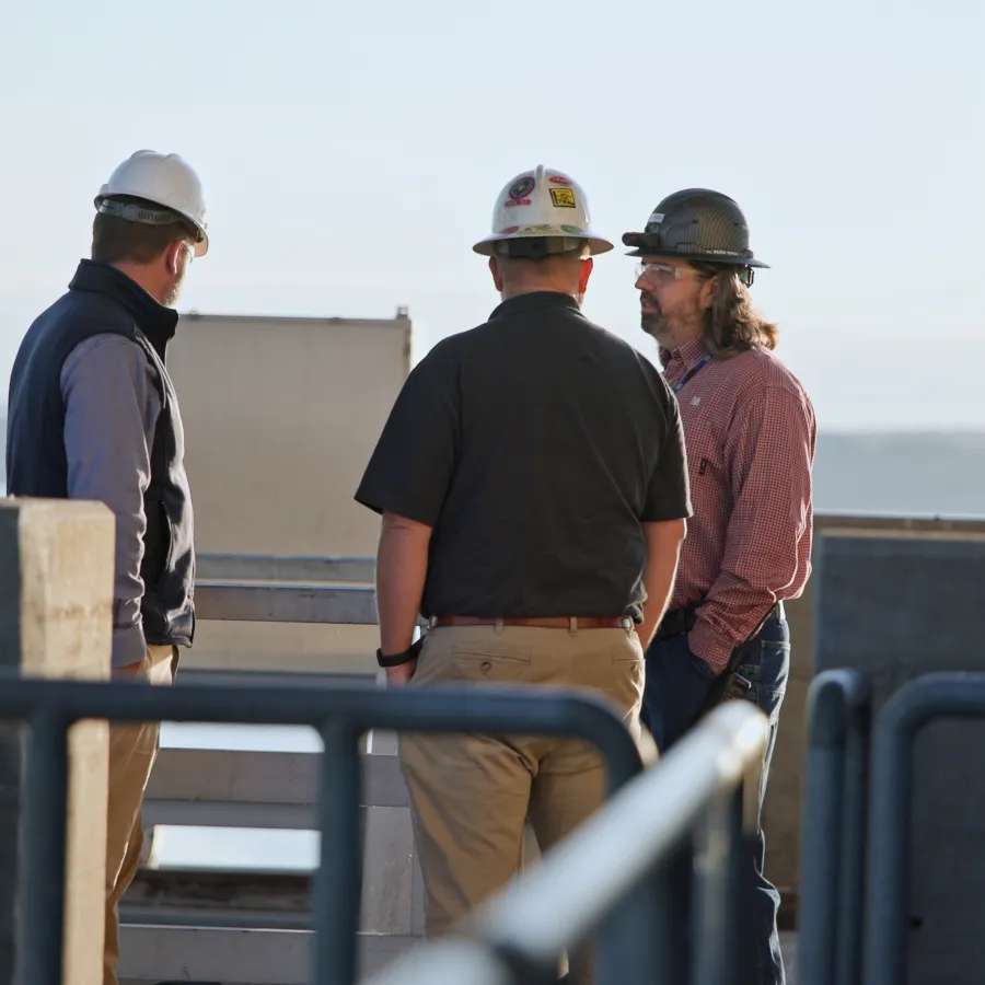 Construction workers wearing hard hats discuss project outdoors near railings with a water view in the background