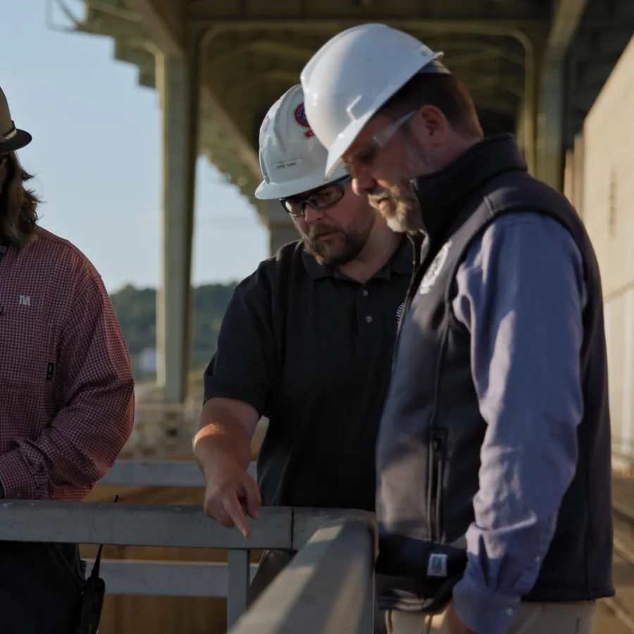 Three construction workers wearing hard hats discuss plans on a metal platform outdoors in daylight.