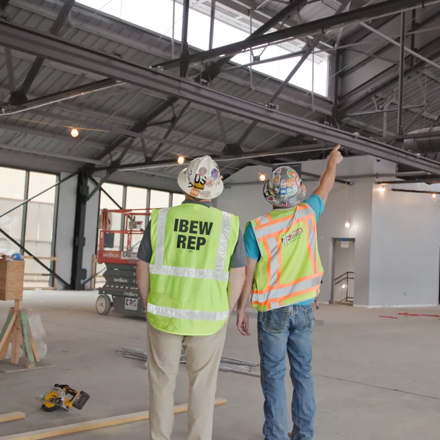 Two construction workers in safety gear inspect and discuss progress inside a large industrial building under construction.