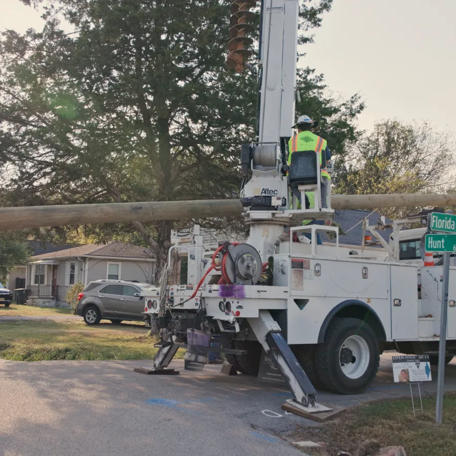 Utility workers installing a large wooden utility pole using a crane truck on a residential street corner.