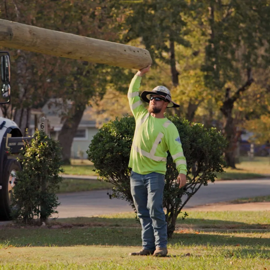 Utility worker in high-visibility clothing carrying a large wooden pole near a utility truck on a suburban street.