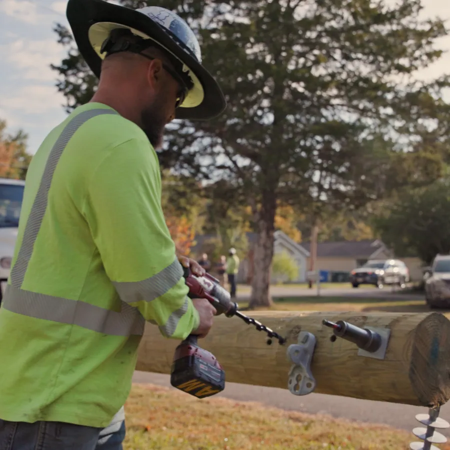 Construction worker in safety gear drilling into a wooden utility pole outdoors near residential area.