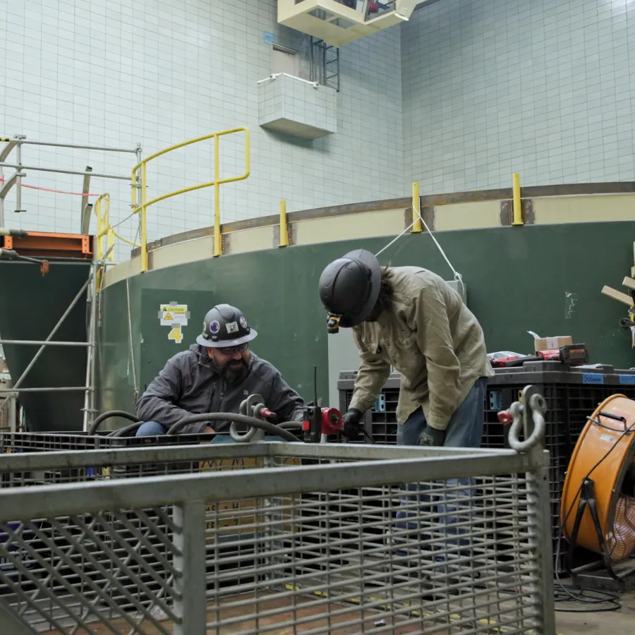 Two workers wearing helmets and protective gear working on industrial equipment in a large factory setting.
