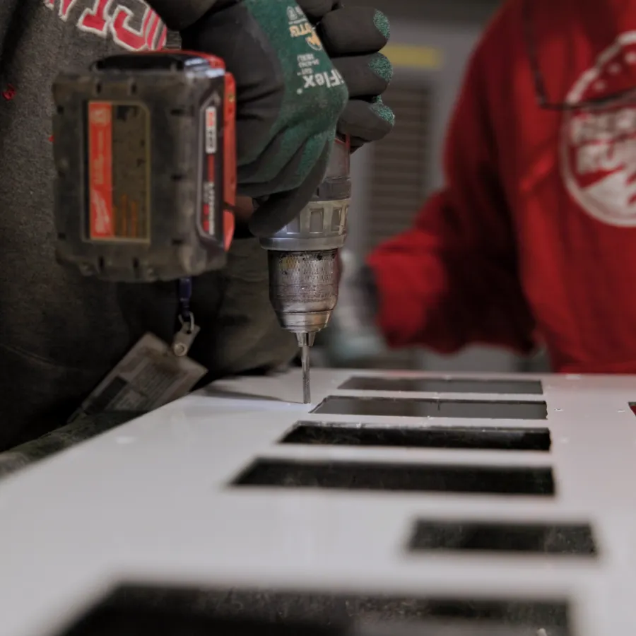 Worker using a power drill on a white panel with rectangular cutouts, another person in red sweatshirt nearby