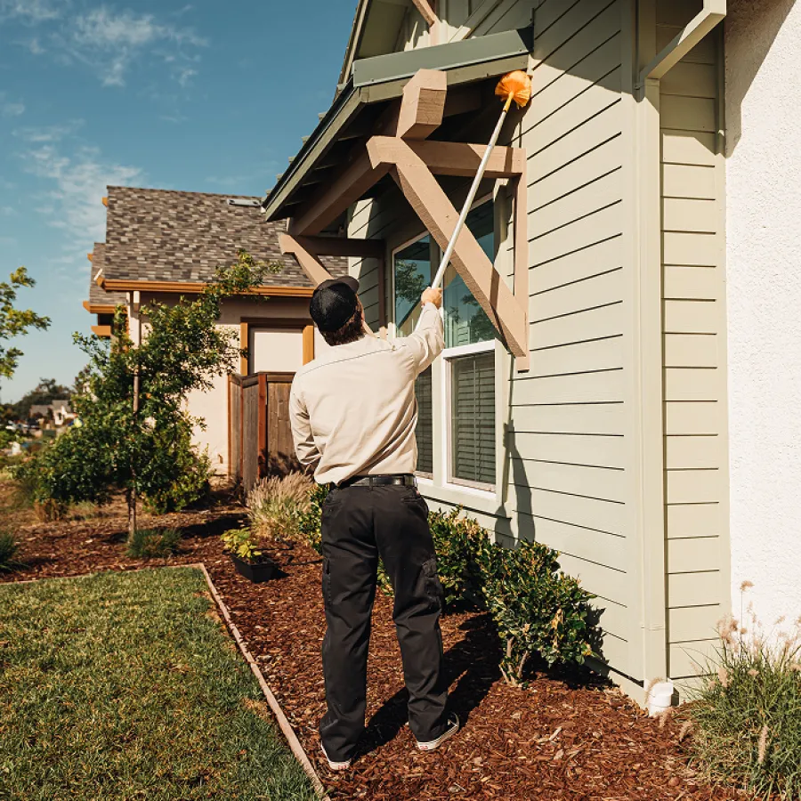 Man cleaning exterior of house with long-handled duster on sunny day in suburban neighborhood