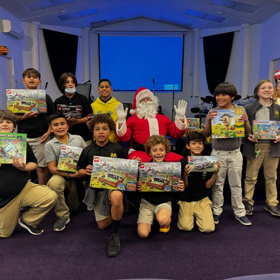 Children and Santa Claus holding LEGO sets inside a community hall with purple carpet and stage.