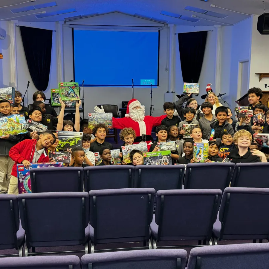 Group of children posing with Santa Claus holding various toy boxes inside a hall with chairs and stage.
