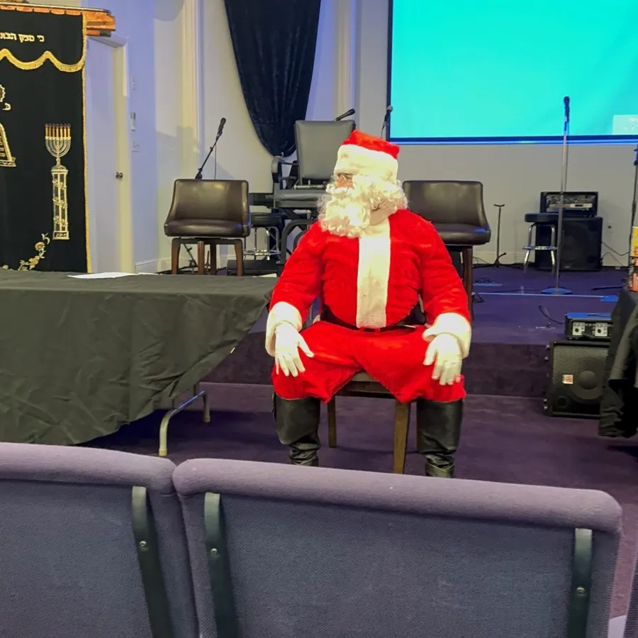 Person dressed as Santa Claus sitting on a chair in a room with chairs, microphone stands, and a projector screen.