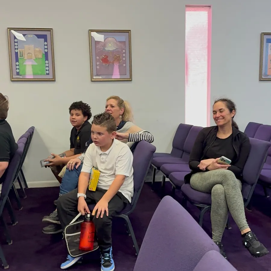 Children and adults seated in a room with purple chairs and framed artwork on white walls.
