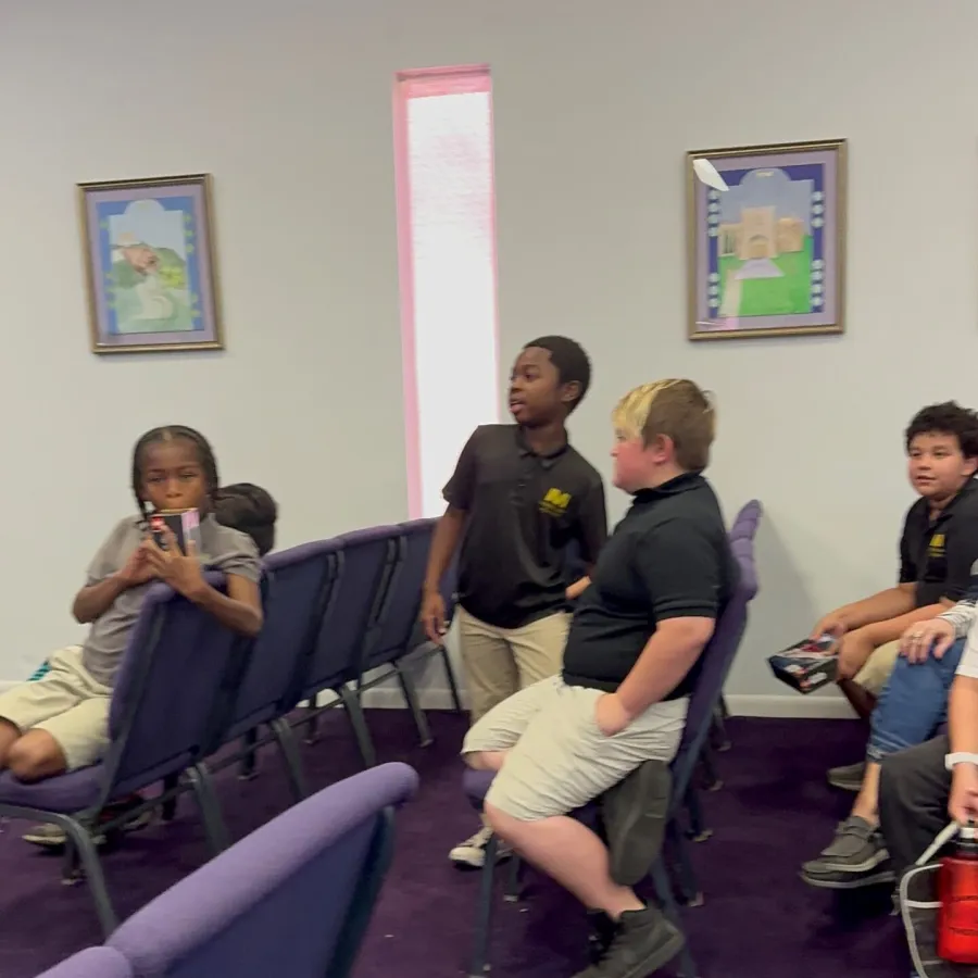 Children in school uniforms sitting and standing between rows of purple chairs in a classroom with framed artwork on the walls.