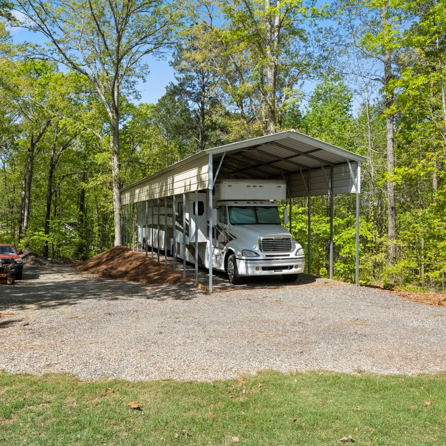 Metal RV carport shelter covering a large white truck surrounded by green trees and gravel driveway.
