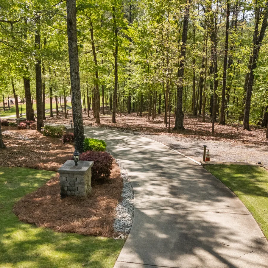 Sunlit curved concrete pathway through a green forest with neatly trimmed grass and stone pillar.