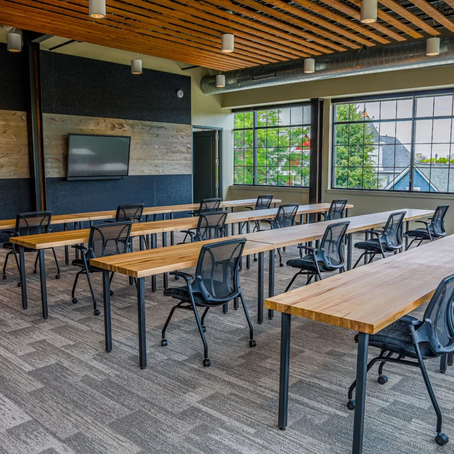 Modern classroom with wooden tables, black mesh chairs, large windows, and ceiling wood paneling lighting.