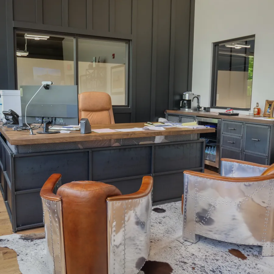 Modern office with metal and leather chairs, large desk, computer, and wooden floors in a bright room.
