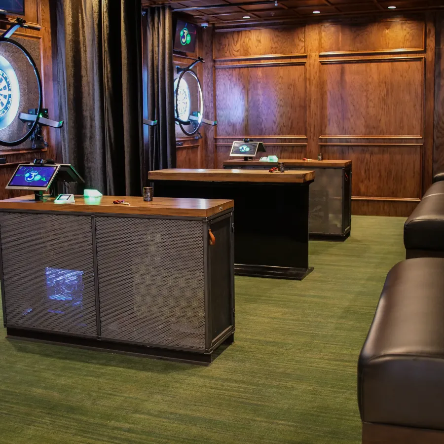 Cozy dart room with wooden accents, two dart boards, interactive screens, and leather seating along the wall