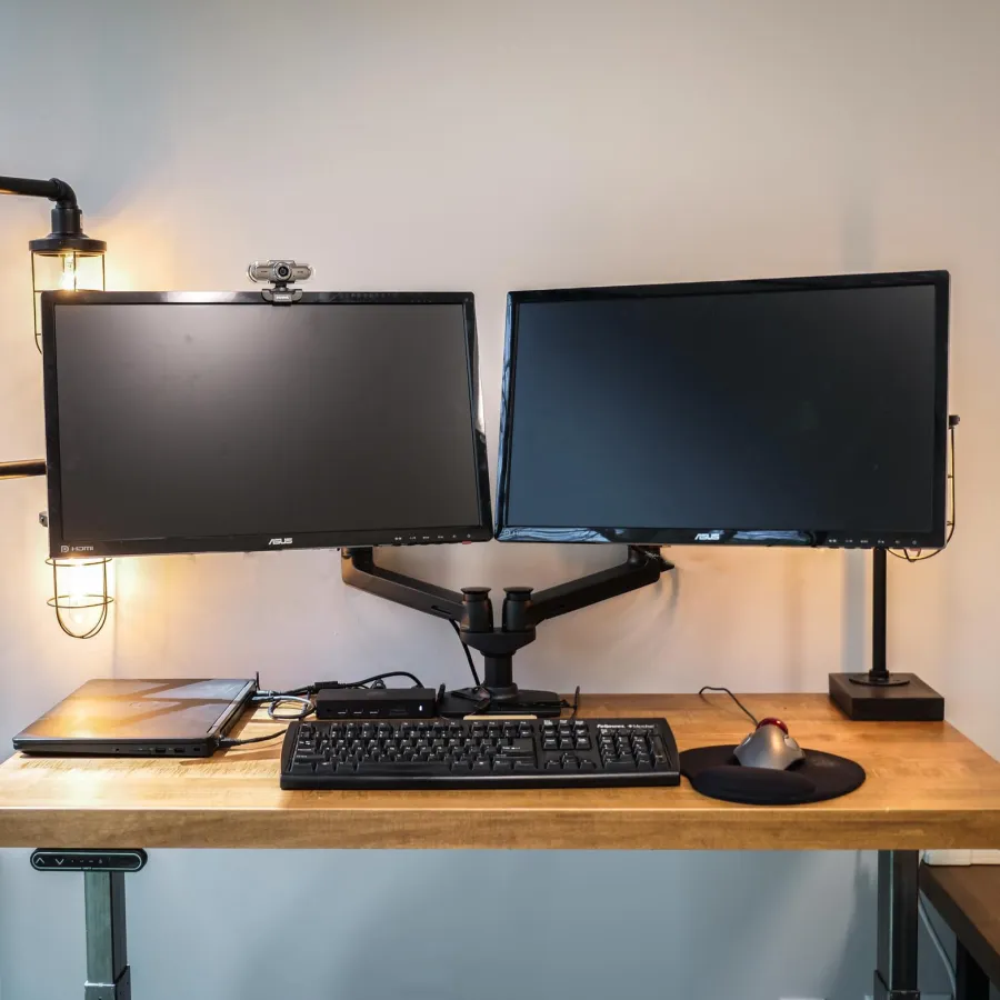 Dual monitors with keyboard and mouse on wooden desk next to laptop and desk lamp against plain wall.