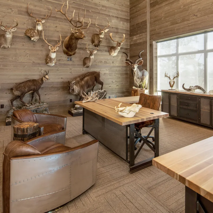 Rustic office with wooden panel walls, mounted animal heads, leather chairs, and wooden desks by a large window