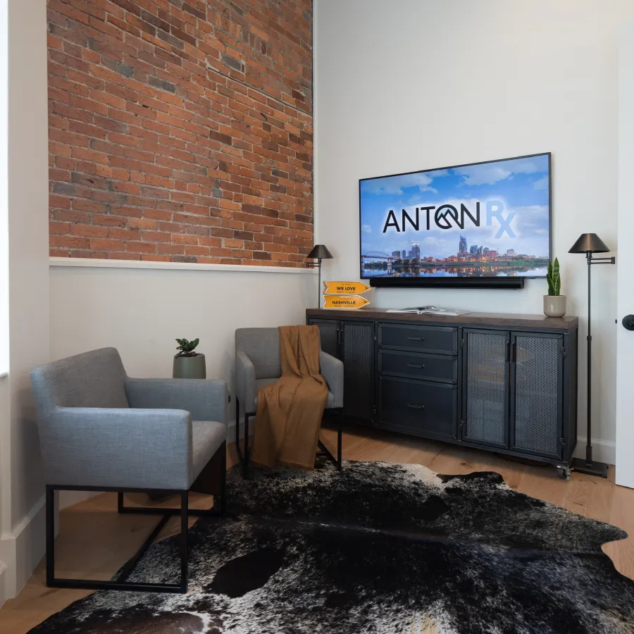 Modern office waiting area with brick wall, gray chairs, black cabinet, TV displaying Anton Rx logo, and floor lamps.