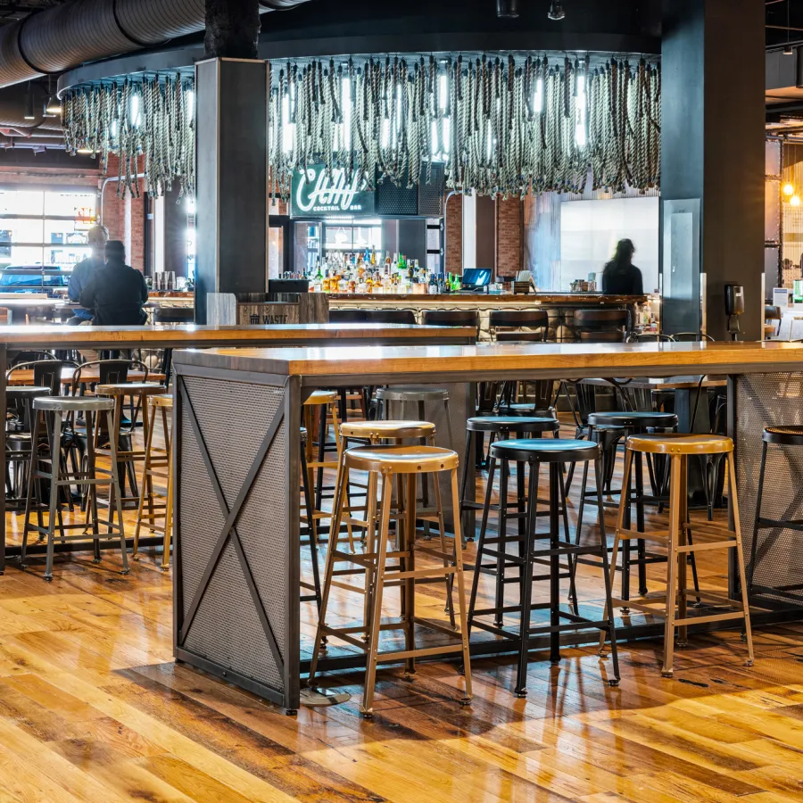 Spacious modern bar interior with wooden floors, high tables, stools, and illuminated decorative ceiling fixtures.