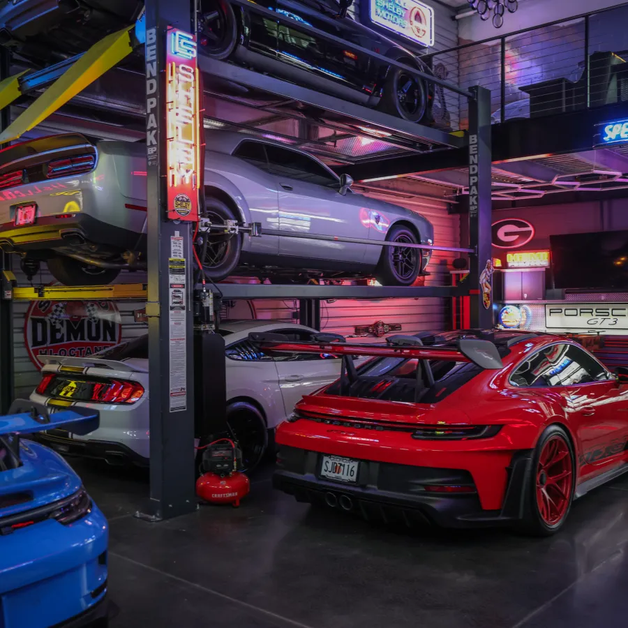 Luxury sports cars in a neon-lit garage with a multi-level car lift and racing paraphernalia.