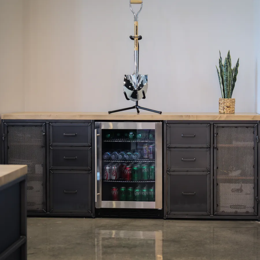 Modern office space with a metal cabinet, a mini fridge stocked with drinks, a decorative shovel, and a potted plant.