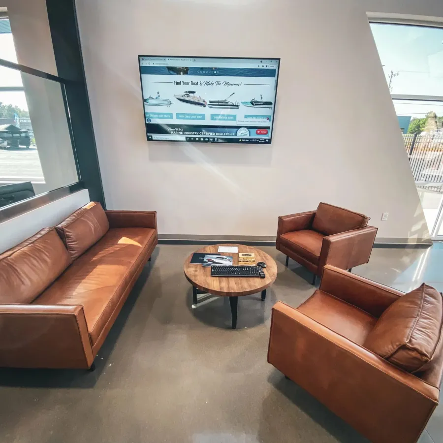 Modern lounge area with brown leather sofa and chairs, round wooden table, and TV screen on wall.