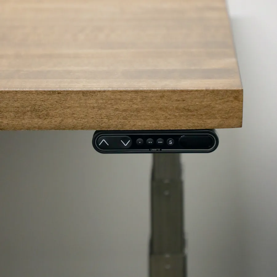 Edge view of wooden adjustable standing desk with control panel and metal leg in neutral background.