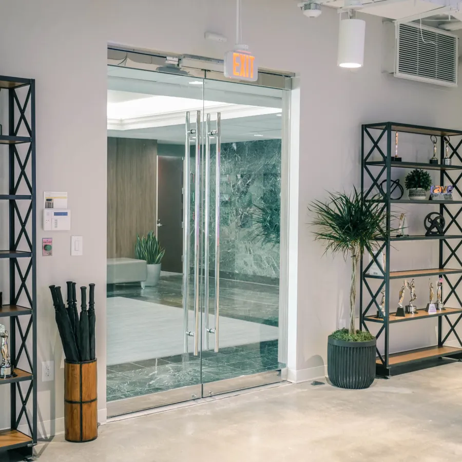 Modern office lobby with glass doors, shelves displaying trophies, black umbrella stand, and potted plants.