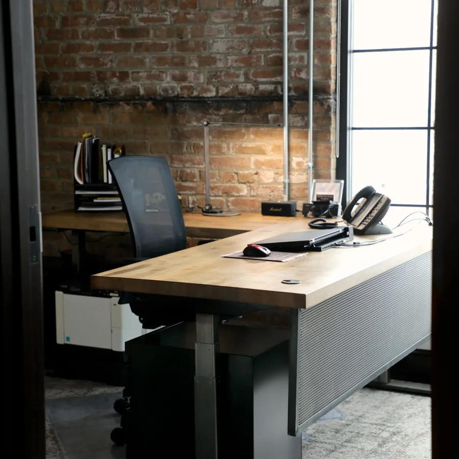 Modern office desk with ergonomic chair, phone, computer mouse, lamp, and brick wall background.