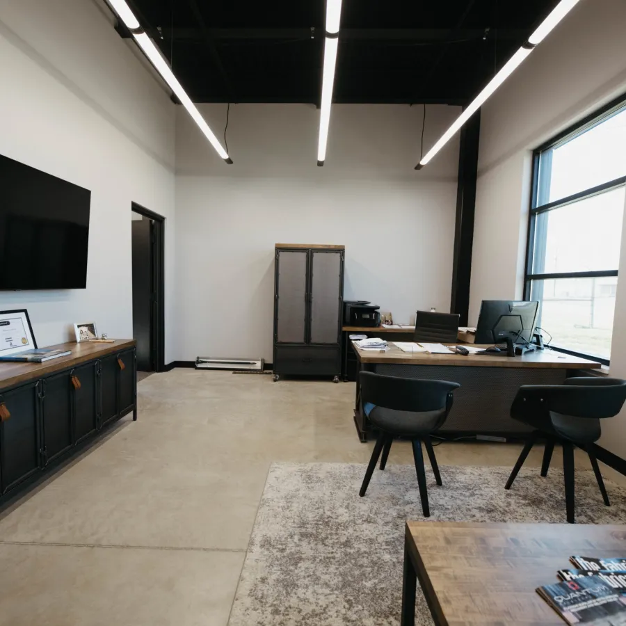 Modern minimalist office with concrete floor, large window, wooden desks, black chairs, and a mounted flat-screen TV.