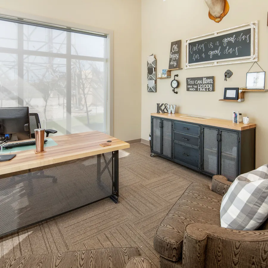 Bright modern office with wooden desk, computer, brown couch, decorative wall art, and large window with blinds.