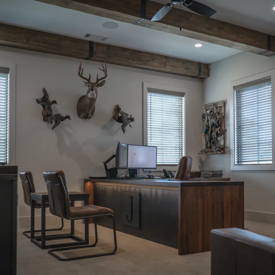 Rustic home office with wooden desk, computer, leather chairs, deer head mount, and blinds on windows