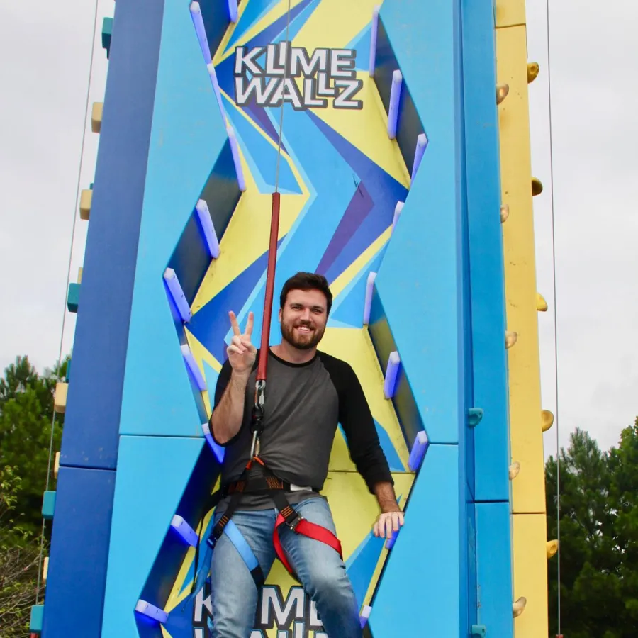 Man wearing harness smiling and posing with a peace sign on a bright blue and yellow climbing wall outdoors.