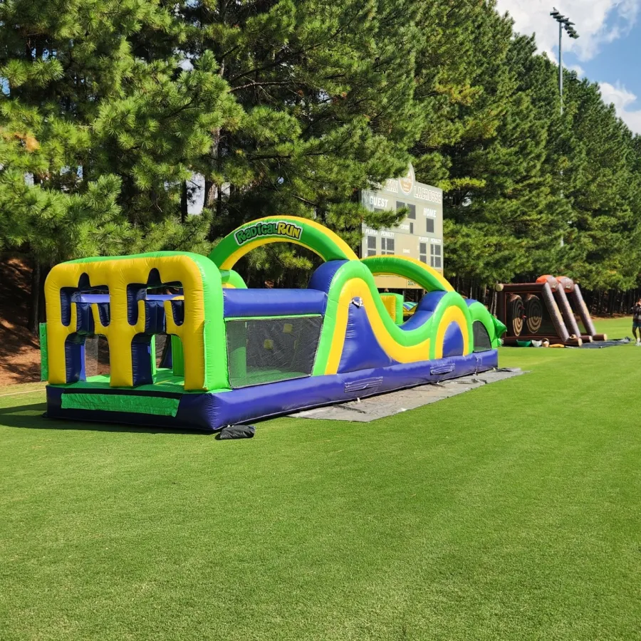Outdoor inflatable obstacle course set up on green grass field with trees and blue sky in background.