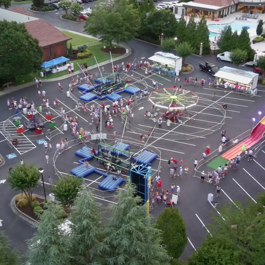 Aerial view of a lively outdoor carnival with rides, games, and people gathered in a parking lot next to buildings.