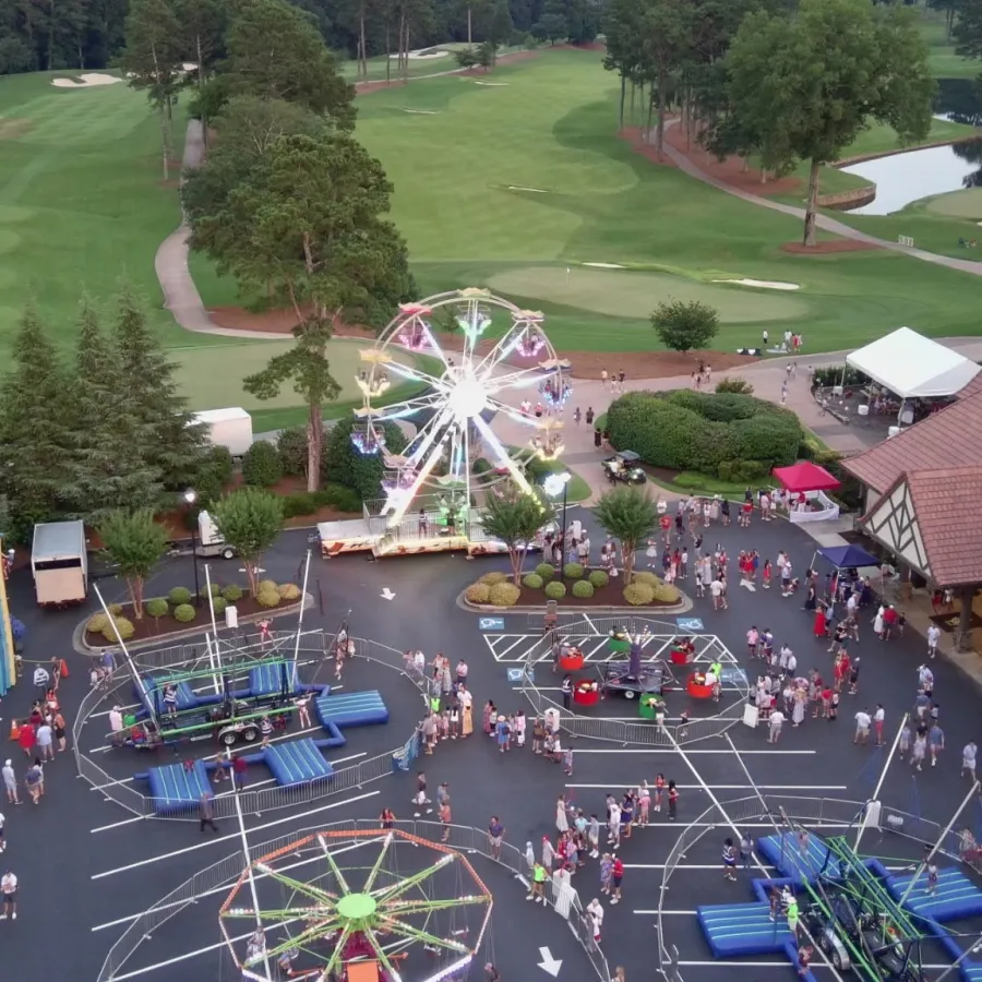 Aerial view of a lively outdoor carnival with rides, crowds, trees, and a golf course in the background at dusk