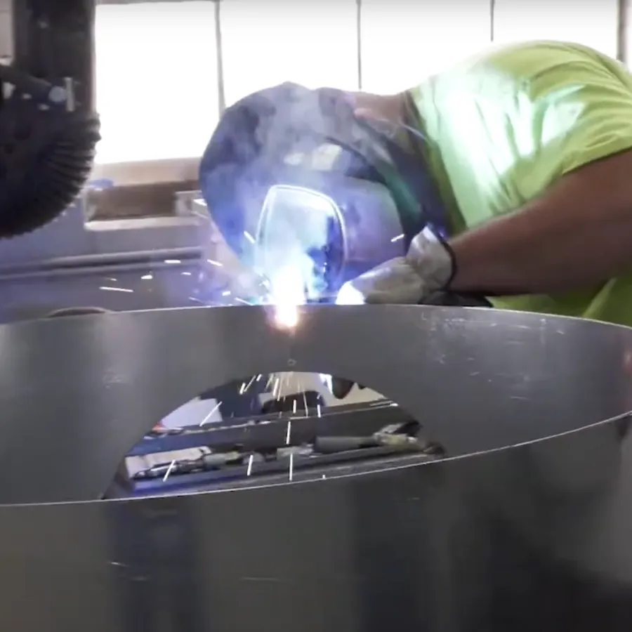 Worker in safety gear welding a large metal cylindrical structure inside a workshop with sparks flying