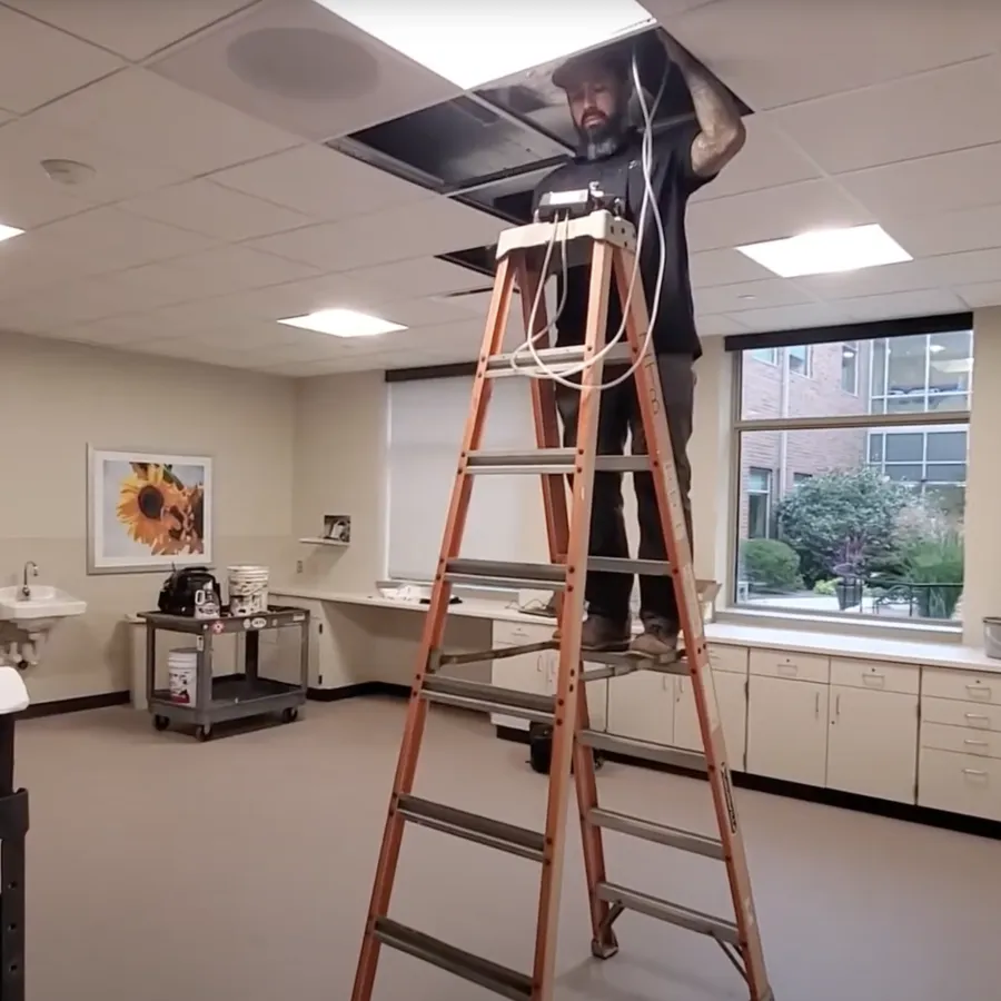 Technician on ladder fixing ceiling panel and electrical wiring in a bright, empty office space with windows.