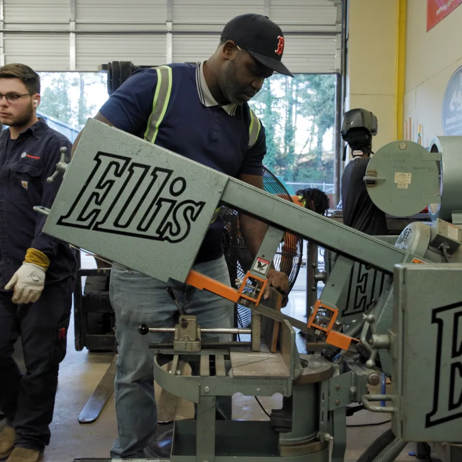 Worker operating Ellis branded metal cutting saw in an industrial workshop with colleagues in the background.