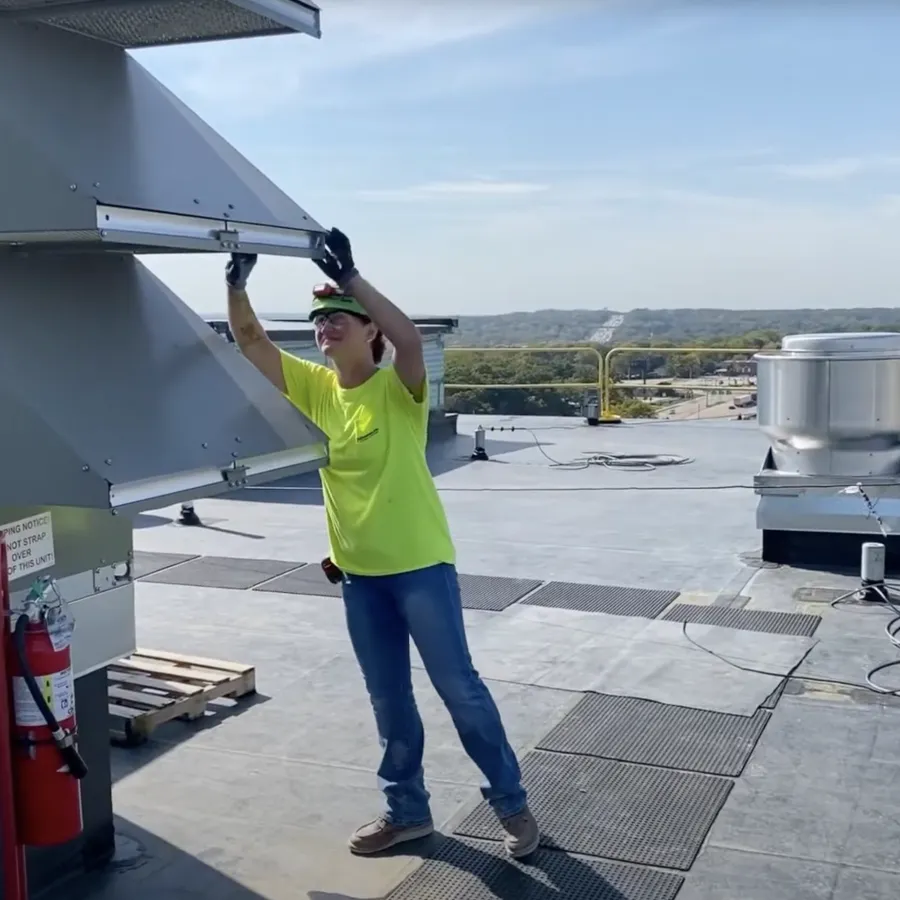 Technician in a neon shirt working on rooftop HVAC equipment under a clear blue sky with safety railing.