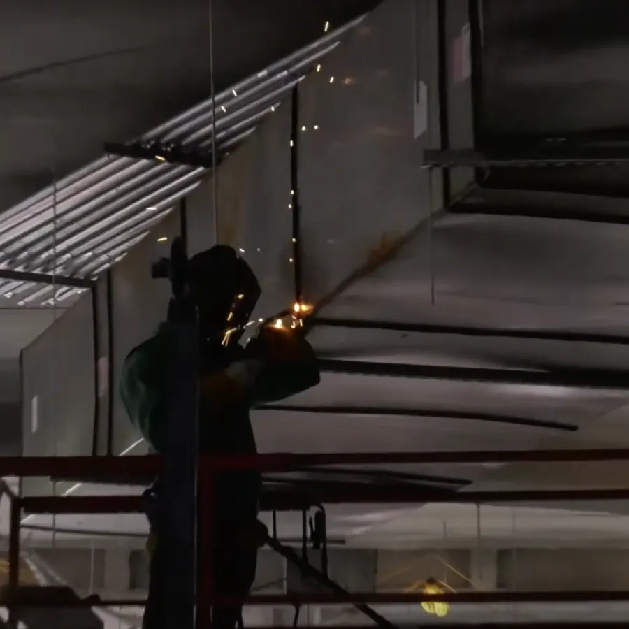 Industrial worker welding metal pipes on elevated platform inside a dimly lit factory.