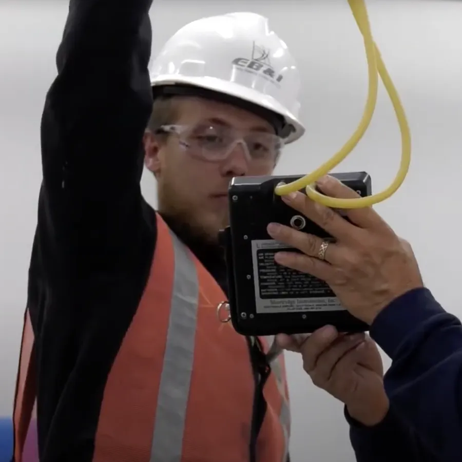 Two construction workers with helmets and safety vests inspecting electrical wiring and a device indoors.